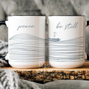 May include: Two white ceramic mugs with black handles. The mug on the left says "peace" and the mug on the right says "be still". Both mugs feature a black and white line drawing of waves with a small boat in the waves on the right mug.