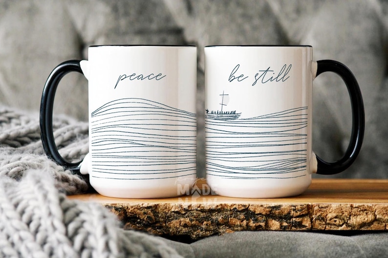 May include: Two white ceramic mugs with black handles. The mug on the left says "peace" and the mug on the right says "be still". Both mugs feature a black and white line drawing of waves with a small boat in the waves on the right mug.