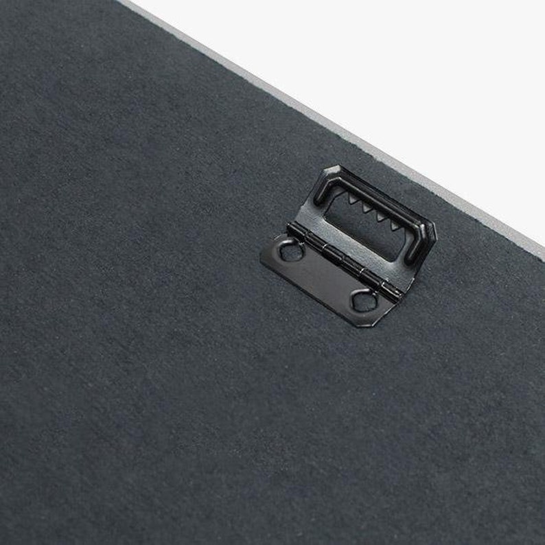 May include: A black metal hanging hook with a sawtooth design on the top and two holes on the sides. The hook is attached to a dark gray surface.