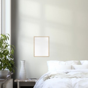 May include: A white bed with white bedding and a white throw blanket. A wooden picture frame hangs on the wall above the bed. A vase with a white textured finish sits on a wooden table next to a potted plant.