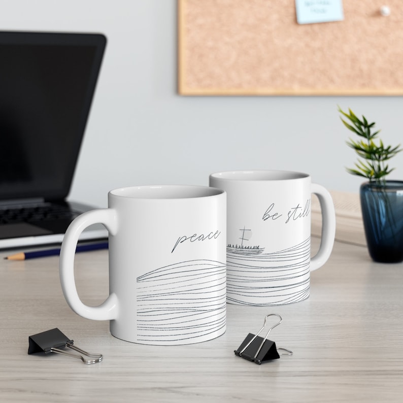 May include: Two white ceramic mugs with black line drawings. One mug says "peace" and the other says "be still". Both mugs feature a stylized drawing of a boat with a cross on the bow.