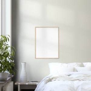 May include: A white bed with white bedding and a white throw blanket. A wooden picture frame hangs on the wall above the bed. A vase with a white design sits on a wooden table next to a green plant.