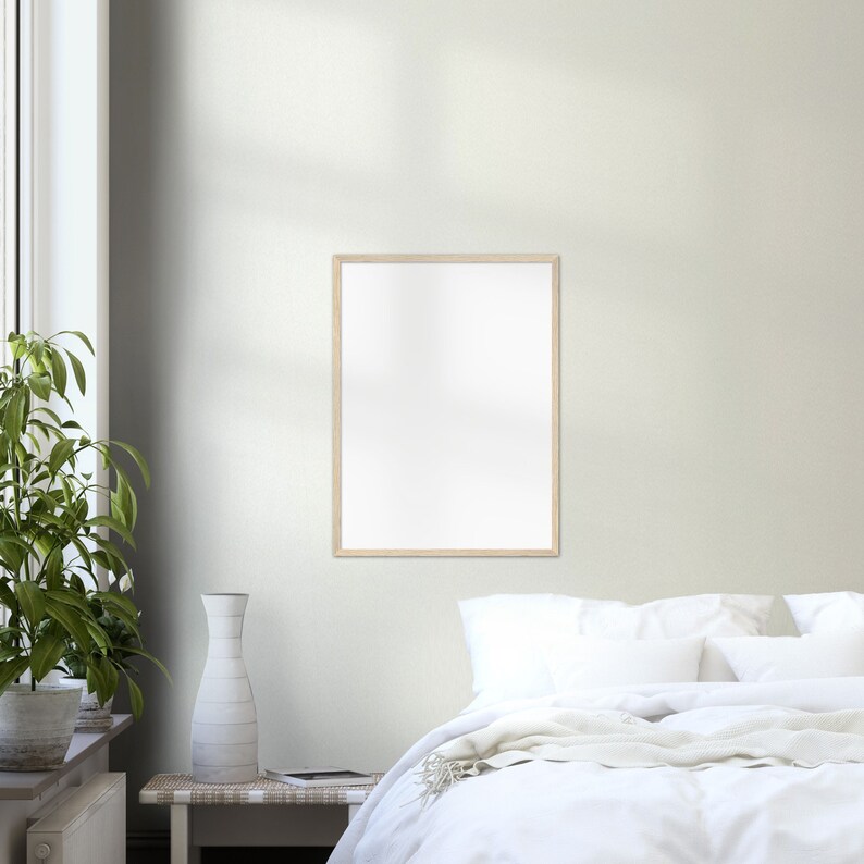 May include: A white bed with white bedding and a white throw blanket. A wooden picture frame hangs on the wall above the bed. A vase with a white design sits on a wooden table next to a green plant.