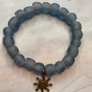 May include: A blue glass bead bracelet with a gold sun charm. The bracelet is made of frosted glass beads and has a simple design.