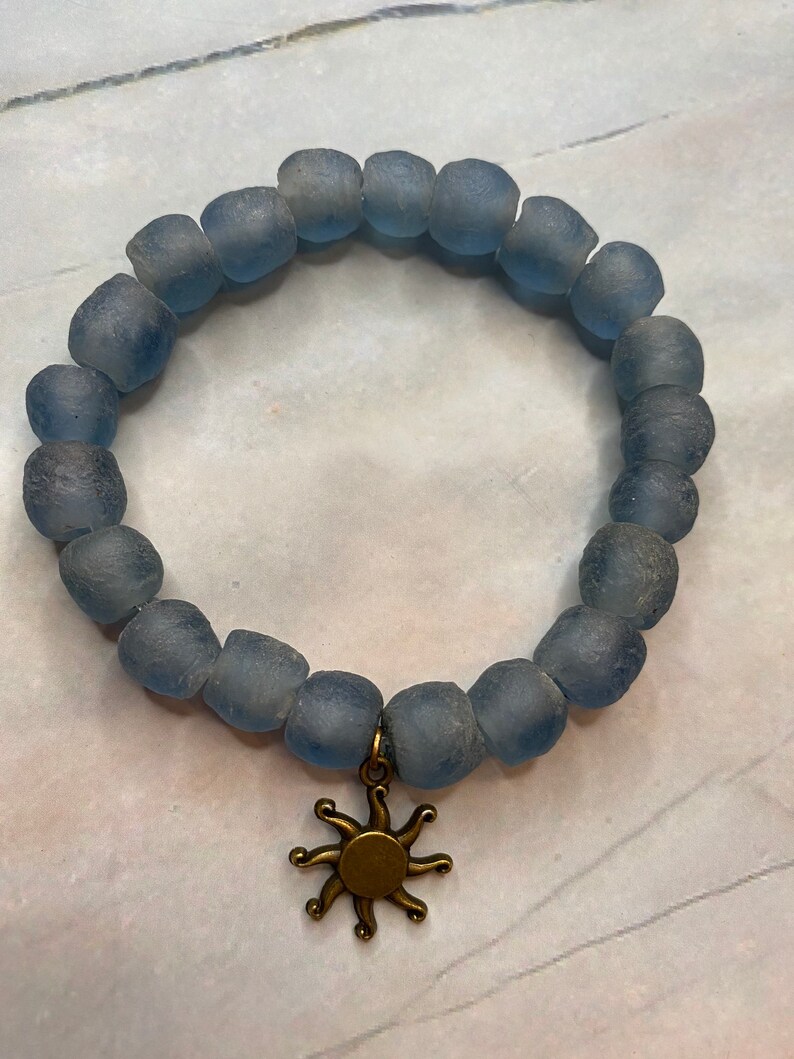 May include: A blue glass bead bracelet with a gold sun charm. The bracelet is made of frosted glass beads and has a simple design.