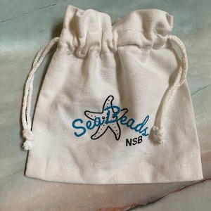 May include: A white cotton drawstring bag with a blue and black logo that says "Sea Beads" and "NSB". The logo features a stylized starfish.