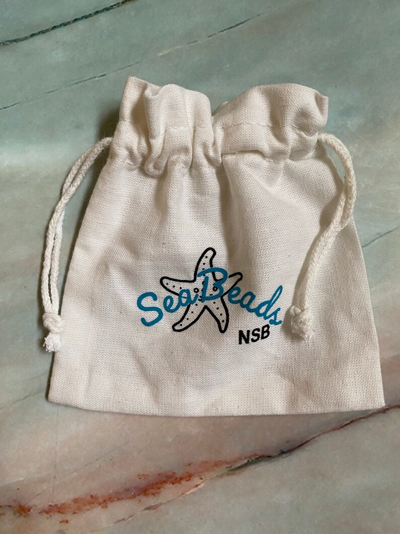 May include: A white cotton drawstring bag with a blue and black logo that says "Sea Beads" and "NSB". The logo features a stylized starfish.