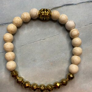 May include: A white and gold beaded bracelet with a decorative gold bead in the center. The bracelet is made of round white beads and faceted gold beads.