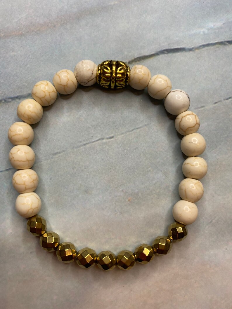 May include: A white and gold beaded bracelet with a decorative gold bead in the center. The bracelet is made of round white beads and faceted gold beads.