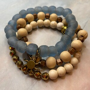 May include: Three layered bracelets with blue, white, and brown beads. The top layer is blue glass beads, the middle layer is white and brown beads, and the bottom layer is gold beads with a sun charm.