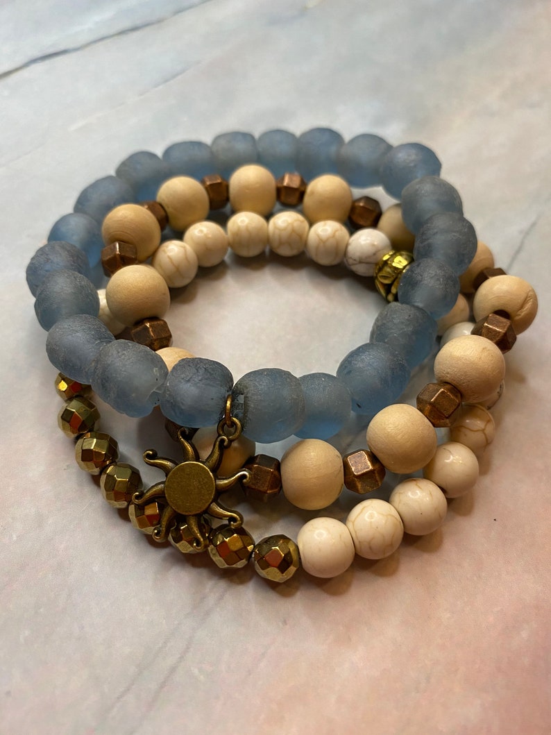 May include: Three layered bracelets with blue, white, and brown beads. The top layer is blue glass beads, the middle layer is white and brown beads, and the bottom layer is gold beads with a sun charm.