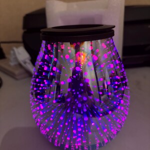 May include: A decorative glass oil warmer with a black rim. The clear glass is adorned with a pattern of pink and purple dots, creating a starburst effect. The warmer is designed to hold scented oils.