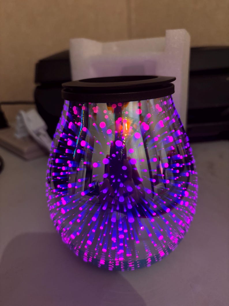 May include: A decorative glass oil warmer with a black rim. The clear glass is adorned with a pattern of pink and purple dots, creating a starburst effect. The warmer is designed to hold scented oils.
