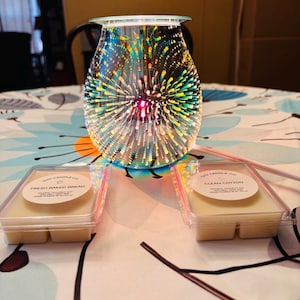 Könnte beinhalten: Ein dekorativer Glas-Wachswärmer mit farbenfrohem Feuerwerksdesign, mit zwei Wachsschmelzbehältern. Ein Behälter ist mit "Fresh Baked Bread" und der andere mit "Clean Cotton" beschriftet.