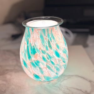 May include: A decorative glass oil warmer with a teal and peach speckled pattern. The warmer has a clear glass top and is illuminated from within, casting a soft glow. The design is reminiscent of sea glass.