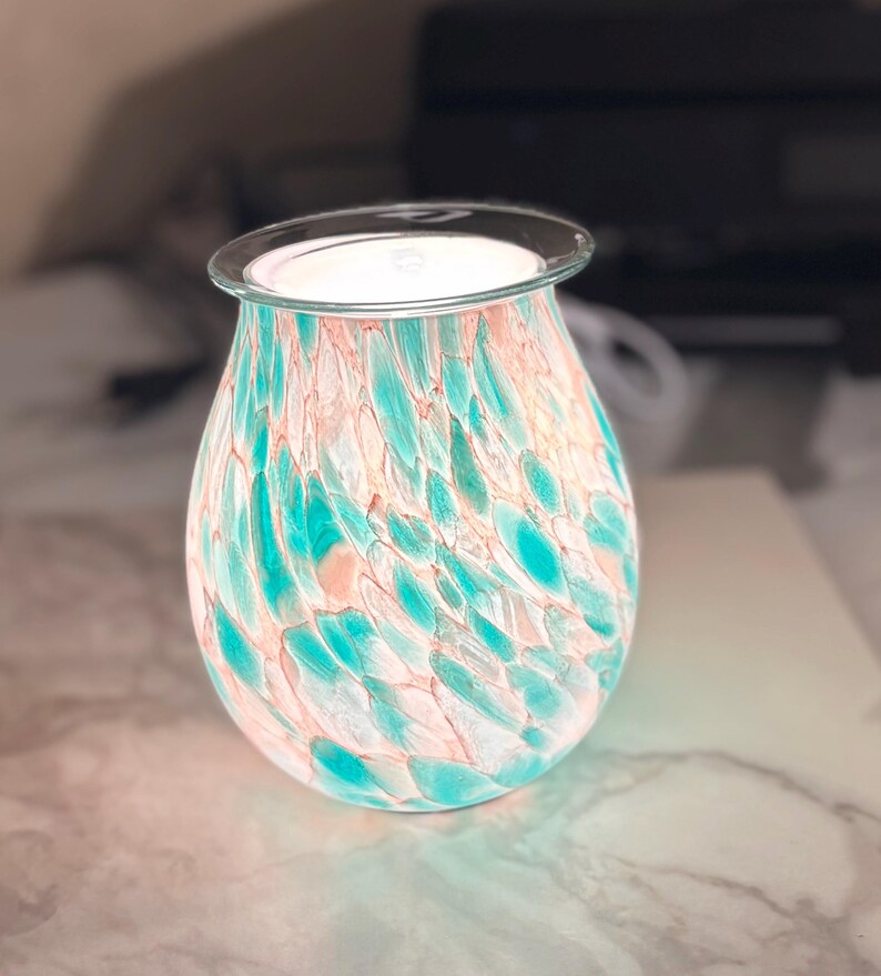 May include: A decorative glass oil warmer with a teal and peach speckled pattern. The warmer has a clear glass top and is illuminated from within, casting a soft glow. The design is reminiscent of sea glass.