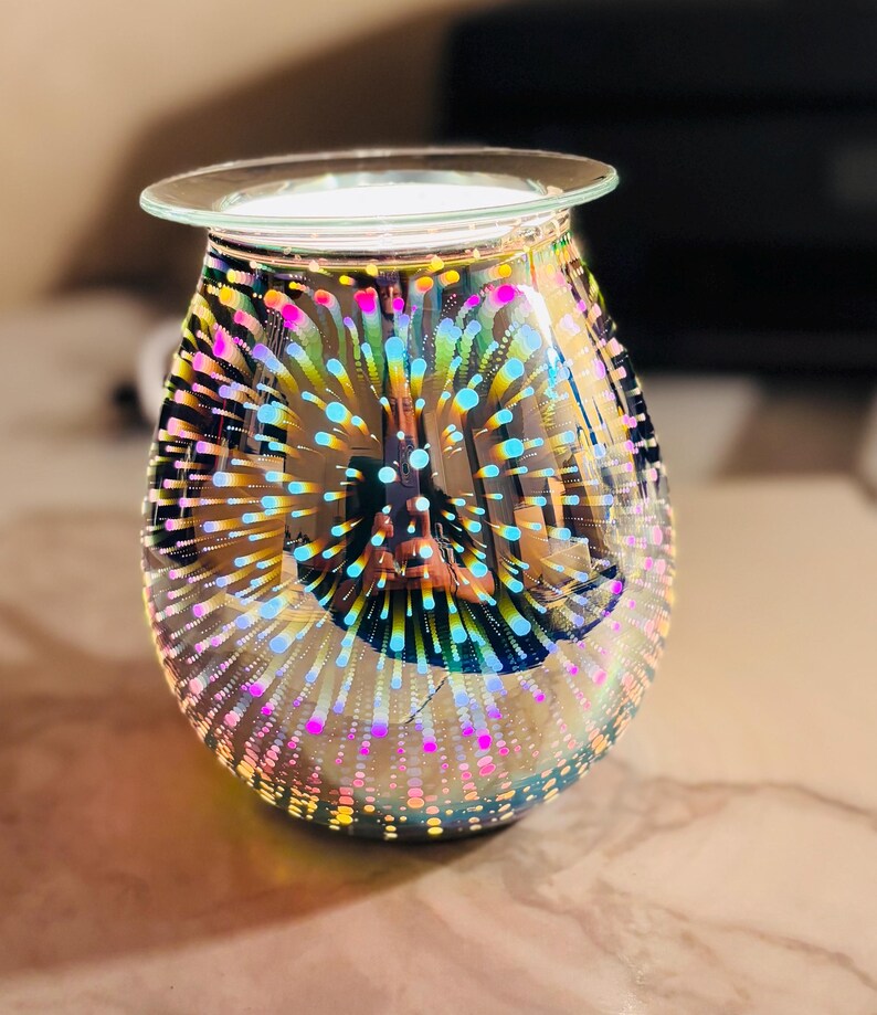May include: A decorative glass oil warmer with a clear, round top and a bulbous body. The glass is iridescent, with a pattern of colorful, radiating dots in shades of pink, blue, yellow, and green. The warmer is designed to hold scented oils.