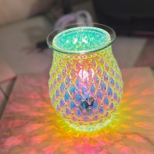 May include: Iridescent glass oil warmer with a textured, scale-like pattern. The top rim is clear glass, and the interior glows with a green light. The base casts colorful light patterns on the surface below.