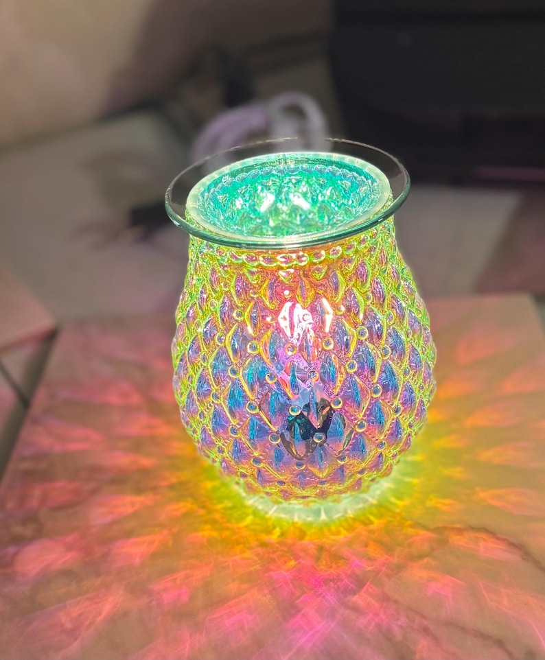 May include: Iridescent glass oil warmer with a textured, scale-like pattern. The top rim is clear glass, and the interior glows with a green light. The base casts colorful light patterns on the surface below.