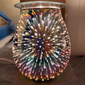 May include: A decorative glass oil warmer with a 3D firework design. The teardrop-shaped warmer features a clear glass dish at the top and a base with a power cord. The design includes colorful bursts of light.