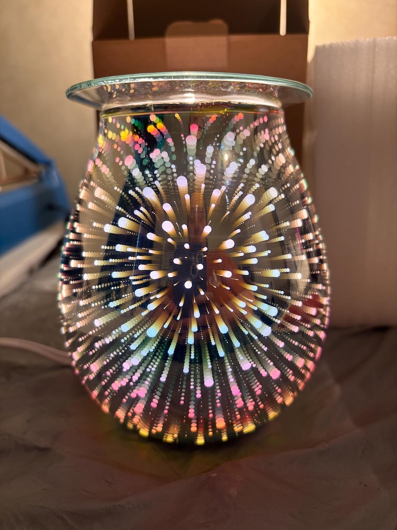 May include: A decorative glass oil warmer with a 3D firework design. The teardrop-shaped warmer features a clear glass dish at the top and a base with a power cord. The design includes colorful bursts of light.