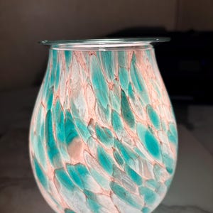 May include: A decorative glass wax warmer with a teal and peach feather-like pattern. The bulbous body of the warmer has a clear glass rim at the top. The interior is illuminated, creating a soft glow.
