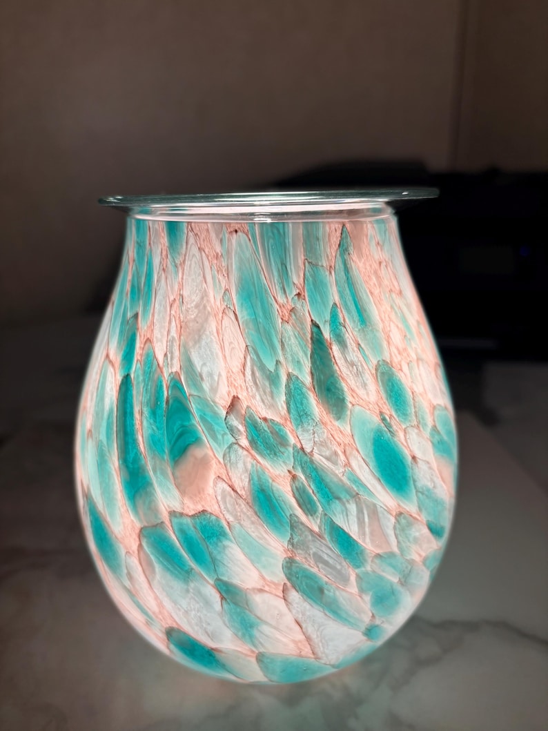 May include: A decorative glass wax warmer with a teal and peach feather-like pattern. The bulbous body of the warmer has a clear glass rim at the top. The interior is illuminated, creating a soft glow.