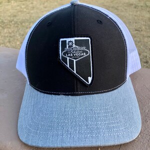 May include: A black, white, and gray trucker hat with a curved brim. The front panel is black with a patch featuring the text "Welcome to Fabulous Las Vegas Nevada". The brim is gray, and the mesh back is white.