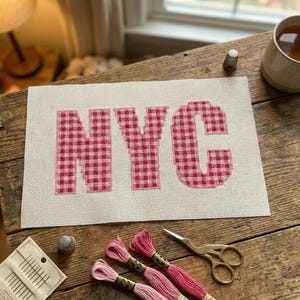 May include: A cross-stitch piece with the letters "NYC" in a red and white gingham pattern. The piece is on a cream-colored fabric, with sewing supplies like thread, needles, and scissors nearby. A thimble and a cup of tea are also visible.
