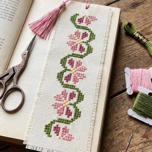 May include: Embroidered bookmark with a pink tassel. The bookmark features a cross-stitch design of pink flowers and green vines on a cream-colored fabric. A pair of scissors, thread, and a needle are also visible.