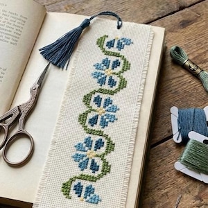 May include: A hand-stitched bookmark with a floral pattern in blue and green thread. The bookmark has a blue tassel and is in an open book. Embroidery thread and scissors are also visible.