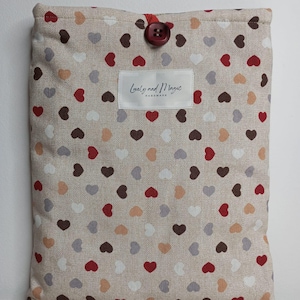 以下が含まれることがあります： 赤いボタン留めで、白いラベルに「Lovely and Magic Handmade」と書かれたベージュのリネンポーチ。ポーチには、赤、茶色、白、桃色の小さなハートの模様が散りばめられています。