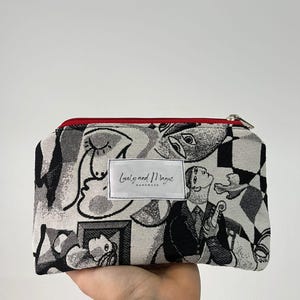 以下が含まれることがあります： 赤色のジッパーが付いた、白黒のパターンでデザインされたジッパー付きポーチ。ポーチには、「Lovely and Magic Handmade」というテキストが書かれた白いラベルが付いています。