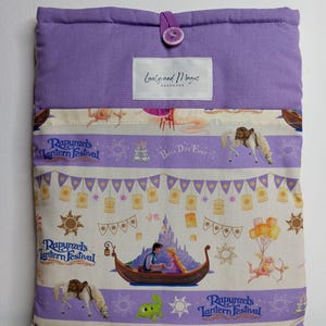 以下が含まれることがあります： ボタン留めの紫色の布製ブックカバー。前面には、ラプンツェルのランタンフェスティバルのプリントが施され、ラプンツェルとフリン・ライダーがボートに乗っているシーンが描かれています。上部のフラップには「Lovely and Magic Handmade」の文字が書かれています。