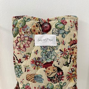 以下が含まれることがあります： ベージュのキルティング生地のケースで、カラフルな花柄とサボテン柄です。ケースには、大きな濃い赤色のボタン留めと、「Lovely and Magic Handmade」と書かれた小さな白いラベルが付いています。