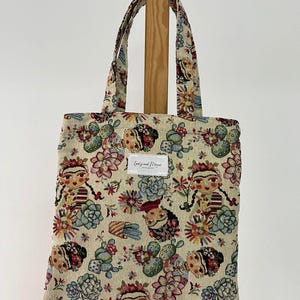 以下が含まれることがあります： ベージュのトートバッグ。カラフルな花柄とフリーダ・カーロにインスパイアされたデザイン。バッグは長方形で、2つのハンドルと「Lovely and Mine」と書かれた小さな長方形のラベルが付いています。デザインにはサボテン、花、肖像画が含まれています。