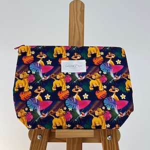Peut inclure: Une pochette zippée bleue avec un imprimé coloré de Simba du Roi Lion. L'imprimé représente Simba, des feuilles de palmier et des fleurs sur un fond bleu foncé. La pochette a une fermeture éclair rouge et une étiquette blanche avec le texte "Cozy and Knot" dessus.