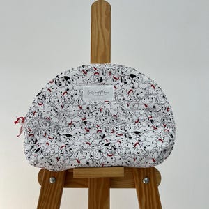 以下が含まれることがあります： 白と黒のダルメシアン柄の布製のメイクアップバッグ。赤と白のラベルには黒い文字で「Crafty and Magic」と書いてあります。バッグは木製イーゼルの上に置かれています。