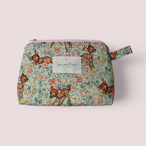 Peut inclure: Une pochette zippée bleu clair avec un imprimé floral représentant Bambi et Panpan. La pochette a une fermeture éclair rose et une anse rose. Le texte "Love and Magic Handmade" est imprimé sur une étiquette blanche.
