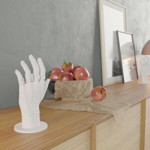 May include: A white hand sculpture with rings on the fingers stands on a wooden surface. A bowl of red apples with green leaves sits on a burlap cloth. Two framed pictures lean against a gray wall.