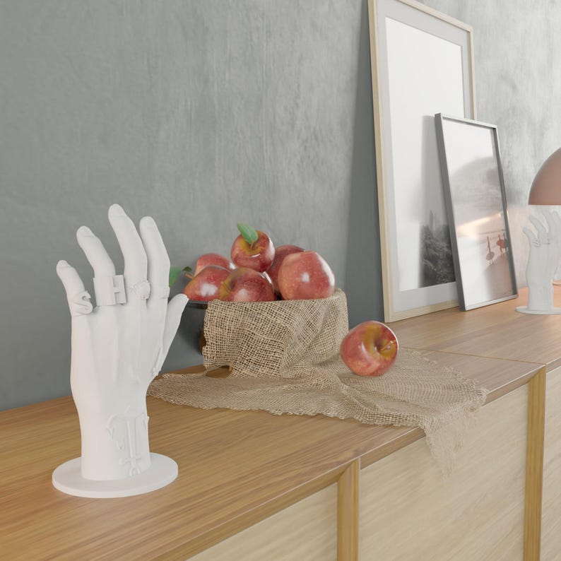 May include: A white hand sculpture with rings on the fingers stands on a wooden surface. A bowl of red apples with green leaves sits on a burlap cloth. Two framed pictures lean against a gray wall.