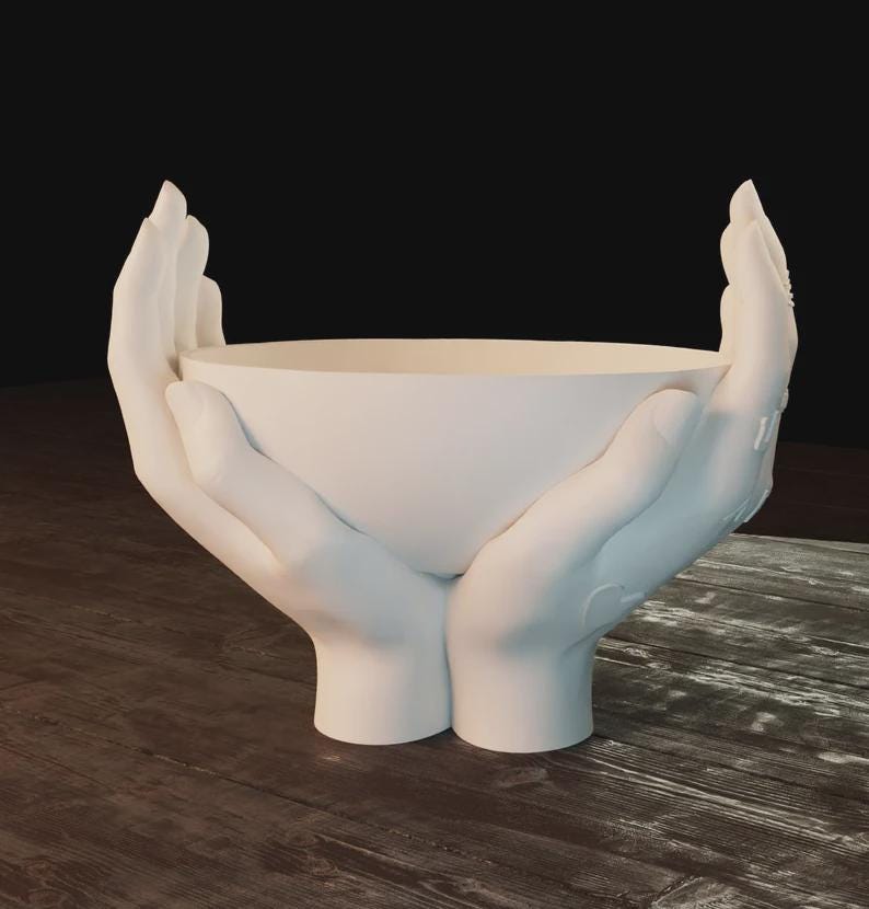 May include: A white ceramic bowl held by two sculpted hands. The hands are positioned as if cradling the bowl, with the fingers gently curved. The bowl and hands are a solid, matte white color.
