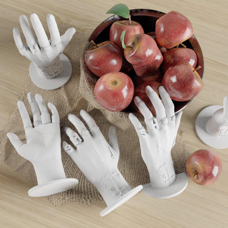 May include: A still-life arrangement featuring white hand-shaped ring displays and a bowl of red apples. The hands are on circular bases, some with rings. The apples are in a dark wooden bowl on a burlap cloth.