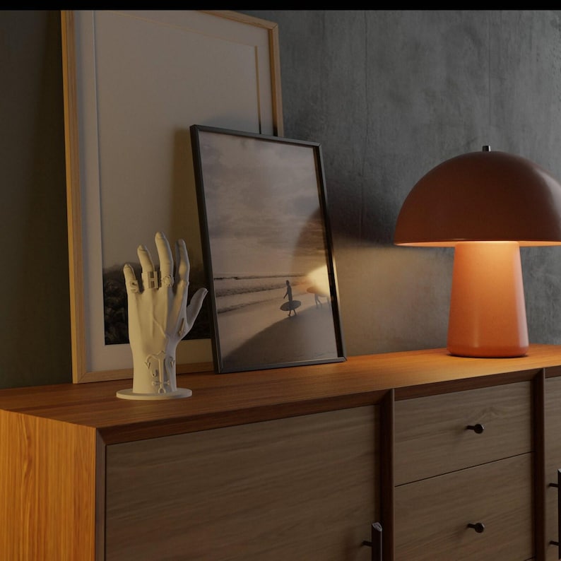 May include: A wooden cabinet with a decorative white hand sculpture, framed artwork, and a modern orange table lamp. The artwork features a beach scene. The lamp has a mushroom-shaped shade.