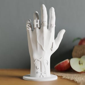 May include: A white, hand-shaped ring holder with rings spelling "SHOE". The holder has a round base and features a stylized design on the palm. The background includes apples and a wooden surface.