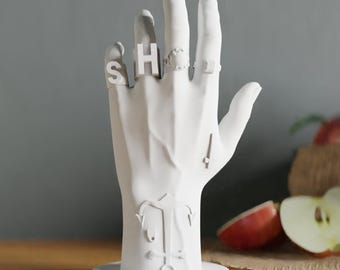 May include: A white, hand-shaped ring holder with rings spelling "SHOE". The holder has a round base and features a stylized design on the palm. The background includes apples and a wooden surface.