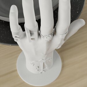 May include: A white hand-shaped jewelry display stand. The stand holds several rings, including one with the letter "S" and another with the letter "H". The base of the stand has a decorative emblem. The rings are silver-colored.