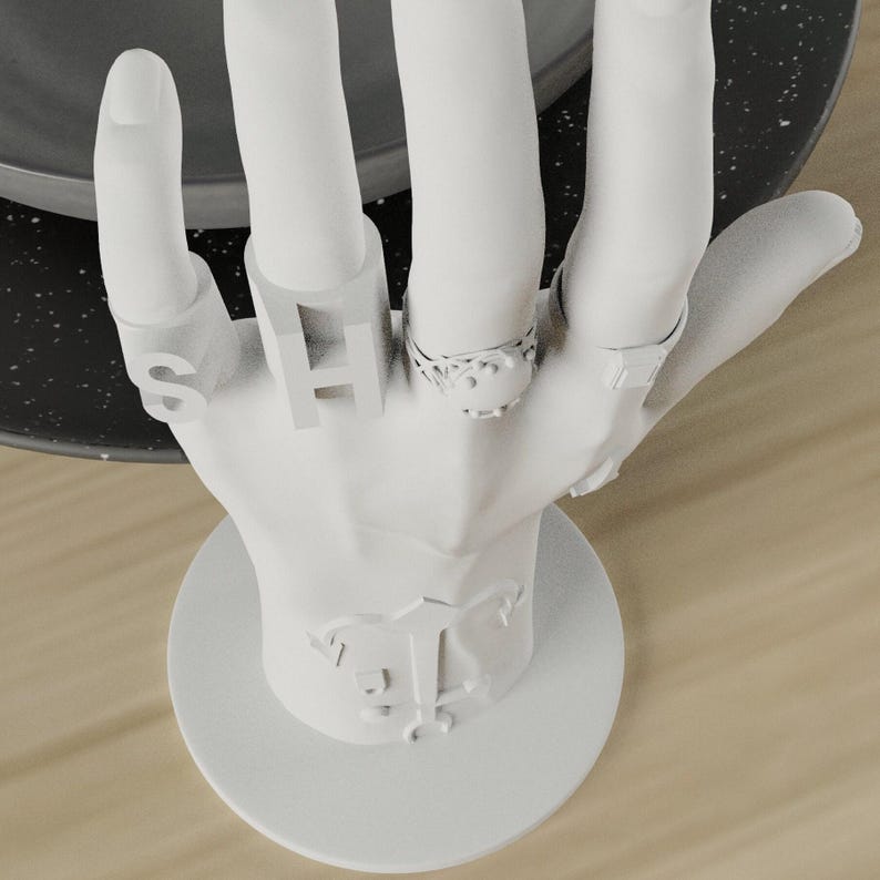 May include: A white hand-shaped jewelry display stand. The stand holds several rings, including one with the letter "S" and another with the letter "H". The base of the stand has a decorative emblem. The rings are silver-colored.