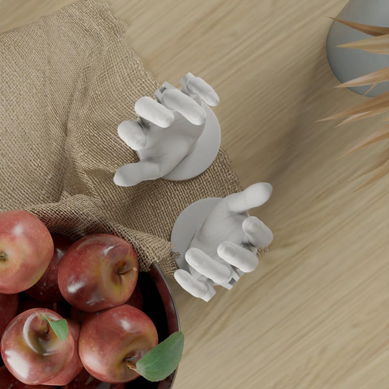 May include: Two white, hand-shaped wall hooks with open palms, positioned near a bowl of red apples. The hooks are mounted on a light brown surface, suggesting a decorative or functional item for a kitchen or dining area.