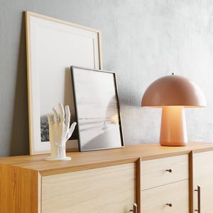 May include: A modern interior scene featuring a wooden cabinet with framed artwork and a mushroom-shaped table lamp. The lamp has a peach-colored shade and base. A white hand sculpture is also on display, adding a touch of artistic flair to the room.